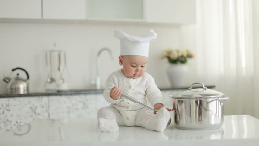 Baby in hat sitting on kitchen table. He holding a soup ladle. Little chef. 
