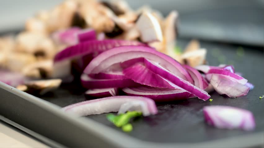 A close-up shot with a narrow focus on freshly sliced red onions on a black cutting board. Chopped mushrooms are softly blurred in the background, ready for cooking.