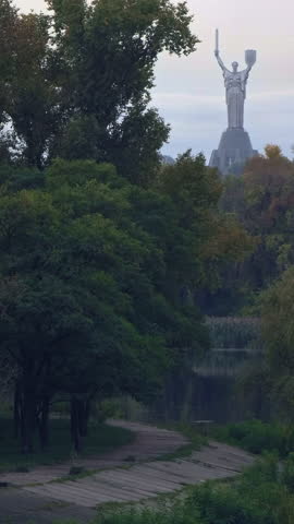 Beautiful view of the Mother Ukraine monument, a national symbol of strength and resilience, standing tall over a tranquil park with lush green trees and a winding pathway alongside a calm river