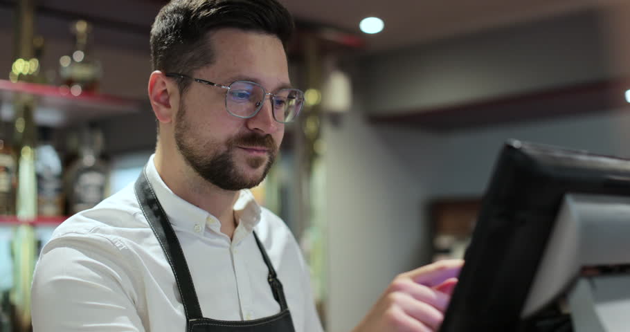 Young waiter with glasses in apron typing on touch screen cash register working at bar or restaurant.