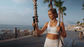 Fitness blogger woman films herself while walking outdoors on a sunny morning. She holds a camera in one hand and carries a yoga mat over her shoulder. Concept of active lifestyle and content creation - Powered by Shutterstock - Get 15% off with code: PIKWIZARD15