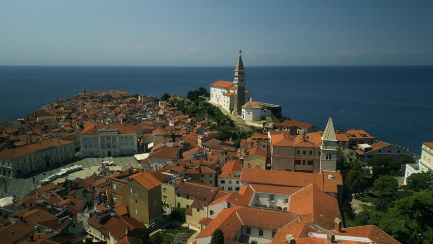 Church stone bell tower rises above old houses with red rooftops on narrow streets. Sea horizon glitters under sunlight surrounding historical European town