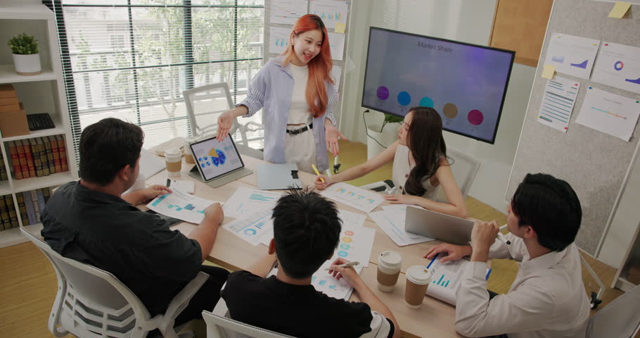 Focused group of young Asian professionals meeting around a table in a modern office. Business team analyzing financial charts, discussing data, and planning corporate project strategy together.