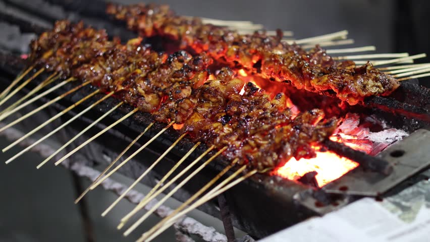 Chicken Satay or Sate Ayam skewers coated in thick peanut sauce, being grilled over glowing charcoal embers on a street vendor