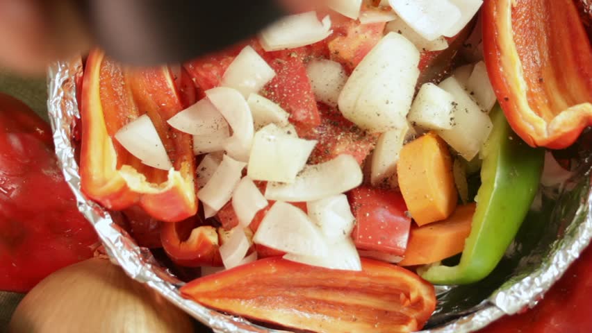 Man sprinkle black pepper from pepper grinder over fresh vegetables in dish with bell pepper, onion, tomato and carrot. Organic food prep and healthy lifestyle cooking in domestic kitchen setting.