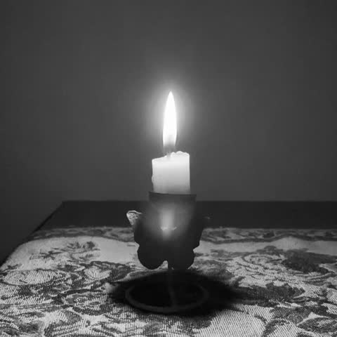 A black-and-white film capturing a bright taper candle glowing upon a butterfly-shaped holder, set before a blue wall and resting on a cloth adorned with floral patterns.