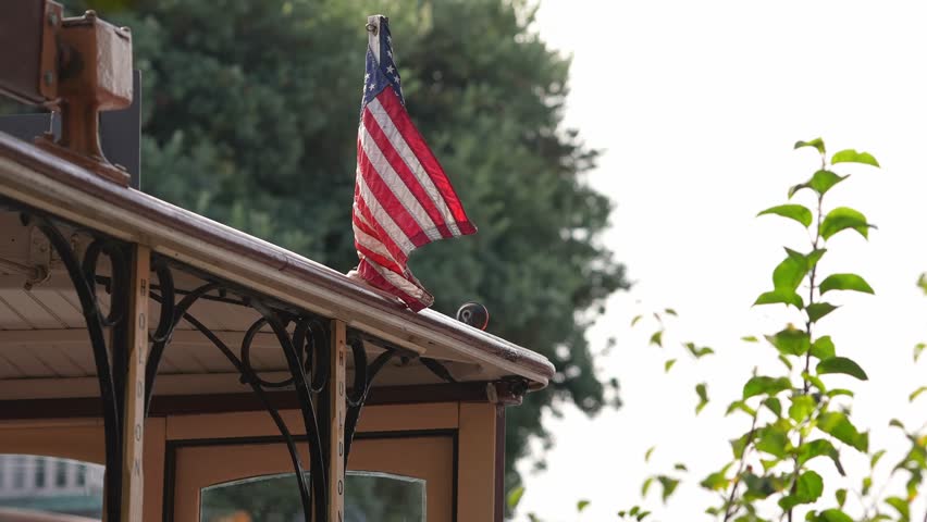 Close daylight shot of a small American flag on a vintage trolley roof in Santa Cruz, California, with gentle motion, shallow depth, backlight, and soft bokeh.