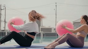 Two focused young women performing a core workout with large pink stability balls outdoors near a swimming pool. Active lifestyle health, group fitness, and strength training in a sunny day morning - Powered by Shutterstock - Get 15% off with code: PIKWIZARD15
