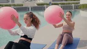 Two focused young women performing a core workout with large pink stability balls outdoors near a swimming pool. Active lifestyle health, group fitness, and strength training in a sunny day morning - Powered by Shutterstock - Get 15% off with code: PIKWIZARD15