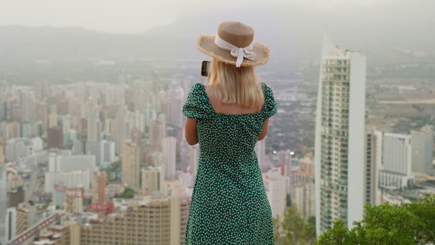 Back view of young blonde woman in green dress and straw hat taking picture of modern cityscape from high viewpoint, enjoy summer vacation and capturing memories of journey.Benidorm Spain