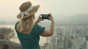 Young woman in straw hat taking photo of coastal city with smartphone from viewpoint, capturing memories of summer vacation travel and sharing them on social media.Technology, blogging, communication  - Powered by Shutterstock - Get 15% off with code: PIKWIZARD15