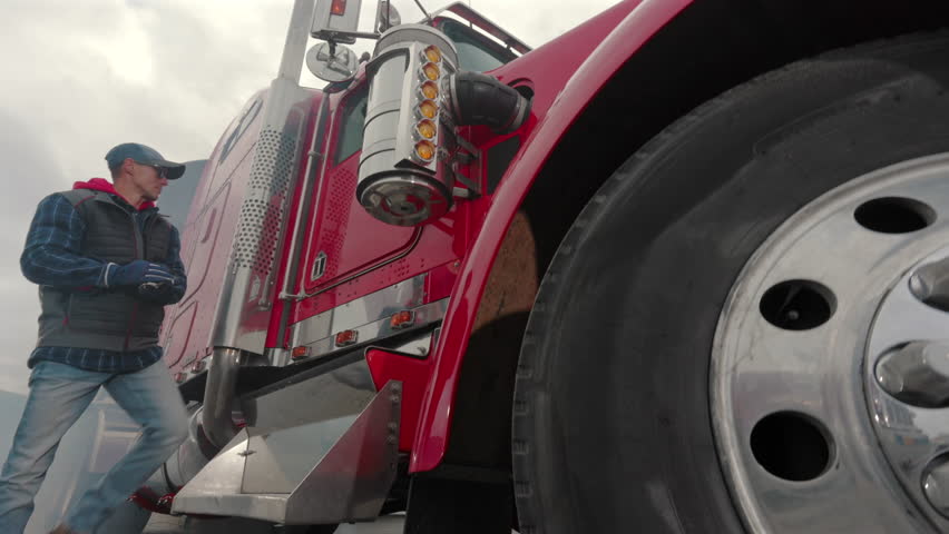 A truck driver inspects the tire and engine of a large red truck in a parking lot. Smoke rises in the background as he assesses the vehicle.
