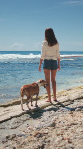 A woman is standing on a sandy beach next to two dogs. Woman is looking out at the ocean while dogs play in the sand. The scene captures a peaceful moment of companionship between human and animals.