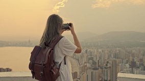 Back view of female tourist with backpack taking picture of urban skyline and sea with mobile phone from high viewpoint during beautiful sunset or sunrise - Powered by Shutterstock - Get 15% off with code: PIKWIZARD15