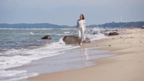 Young woman in light dress walks barefoot along sandy Baltic Sea Beach. Calm sea and scattered rocks create serene atmosphere under soft daylight, with hazy hills in background - Powered by Shutterstock - Get 15% off with code: PIKWIZARD15