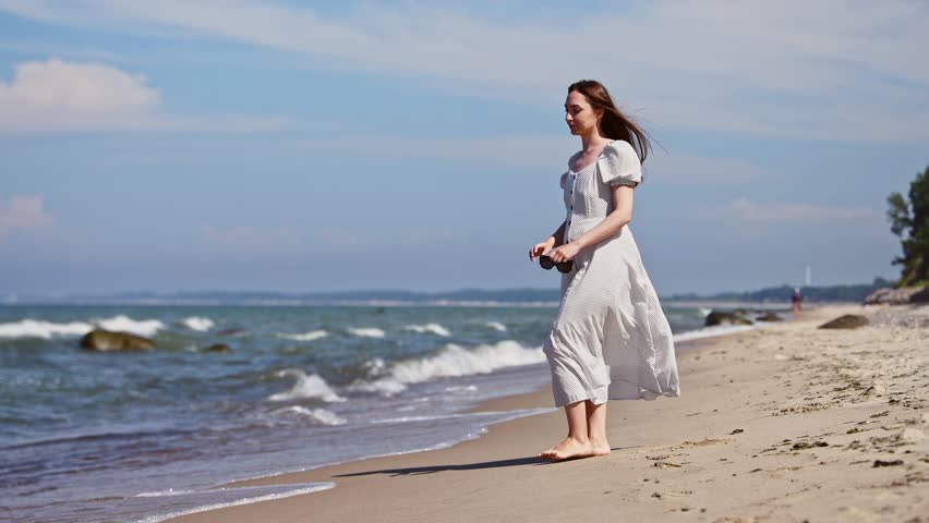 Young woman in flowing white dress and sunglasses stands barefoot on sandy beach of Baltic Sea. Gentle waves lap shore under blue sky with soft clouds, capturing serene coastal moment