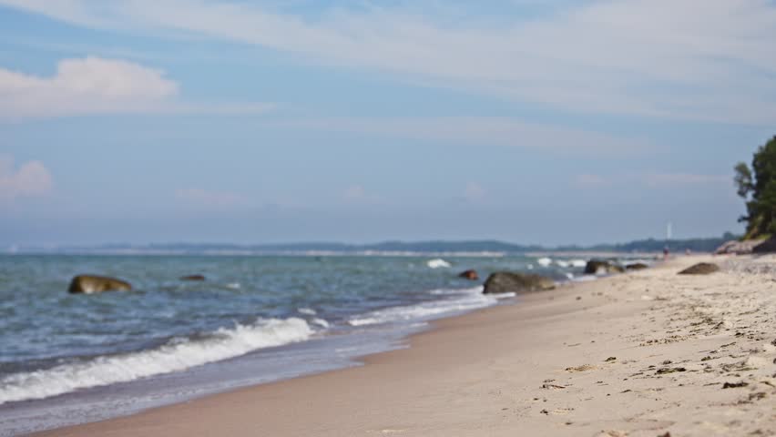 Young woman with long brown hair in white dress stands barefoot on sandy shore, gazing at gentle waves of Baltic Sea under blue sky with scattered clouds
