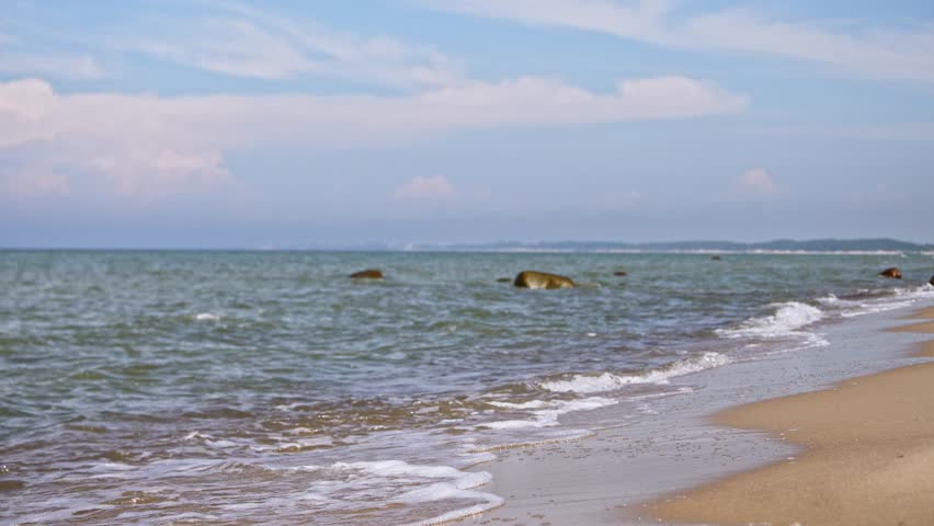 Young woman in white dress walks barefoot at Baltic Seas sandy shore. Gentle waves lap at her feet under bright blue sky with scattered clouds, creating peaceful coastal scene
