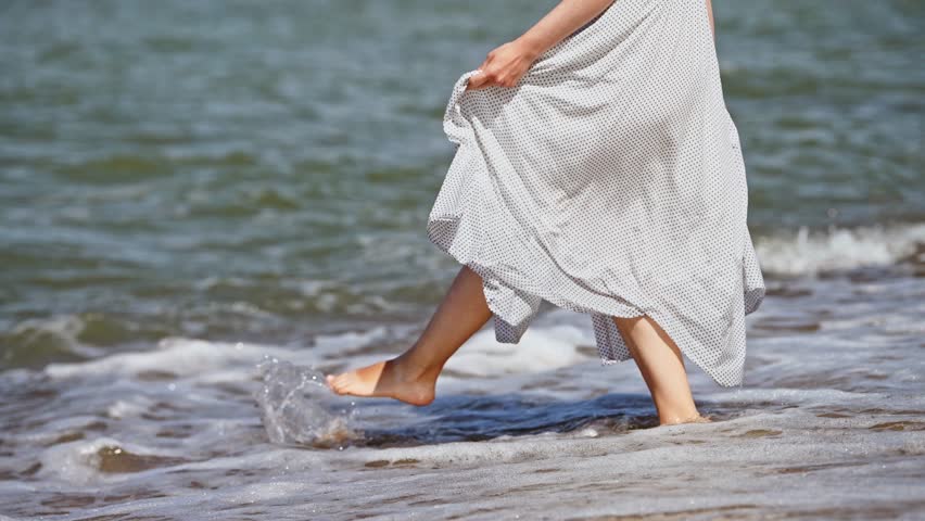 Woman in white polka dot dress stands barefoot in shallow water of Baltic Sea. Gentle waves lap around her feet on sandy shore under natural daylight