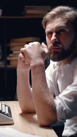 A man sits at his desk, deeply frustrated while trying to write. His dark expression shows the weight of his thoughts. The atmosphere is tense as he battles his inner demons.