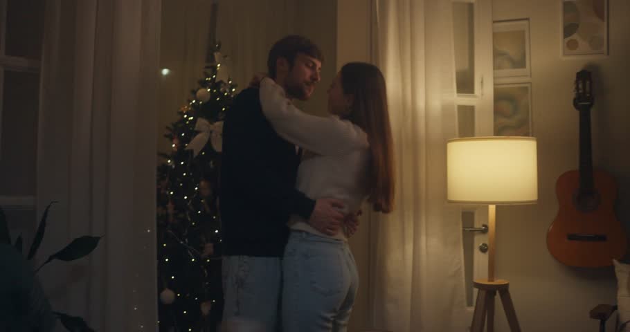 Romantic couple shares a tender moment in a cozy, decorated living room near a Christmas tree in the evening glow