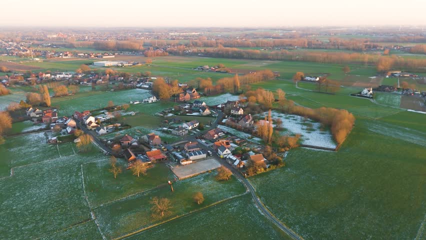 village opwijk belgium yellow trees green fields snow covered rooftops touched by sunlight patches during golden hour under cloudy long view snow-covered