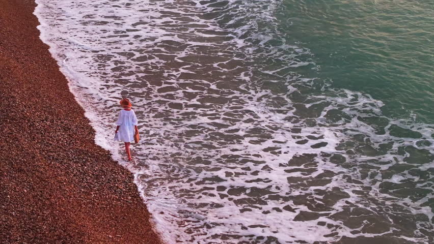 Middle-aged woman in a white dress and straw hat walking barefoot along the seashore at sunrise, carrying a beach bag and enjoying the calm of the morning. High quality 4k footage