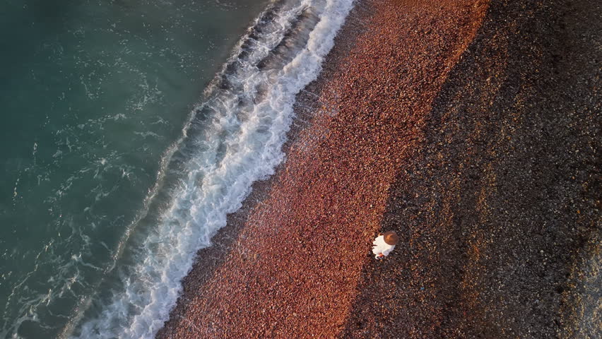 Aerial view of a middle-aged woman in a white dress and straw hat walking alone along a pebbled beach at sunrise, with soft waves touching the shore. High quality 4k footage
