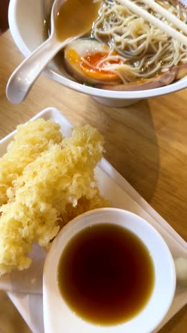 Hand dips crispy shrimp tempura into dipping sauce, ramen bowl visible, bright natural lighting