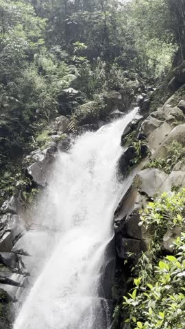 A scenic view of a natural waterfall flowing through rocky cliffs in a tropical forest. The powerful stream of water surrounded by lush greenery showcases the raw beauty of nature in a peaceful