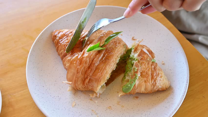 Someone cutting a piece of Matcha (or green tea) croissant for eating. Croissant is a French buttery, flaky and crescent-shaped bread.