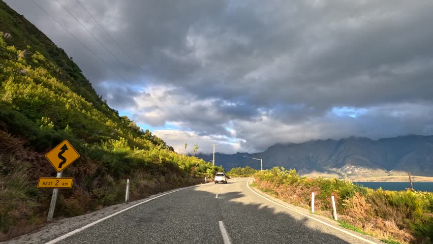 Car travels winding lakeside mountain road under dramatic clouds, late afternoon sunlight, forward-facing perspective