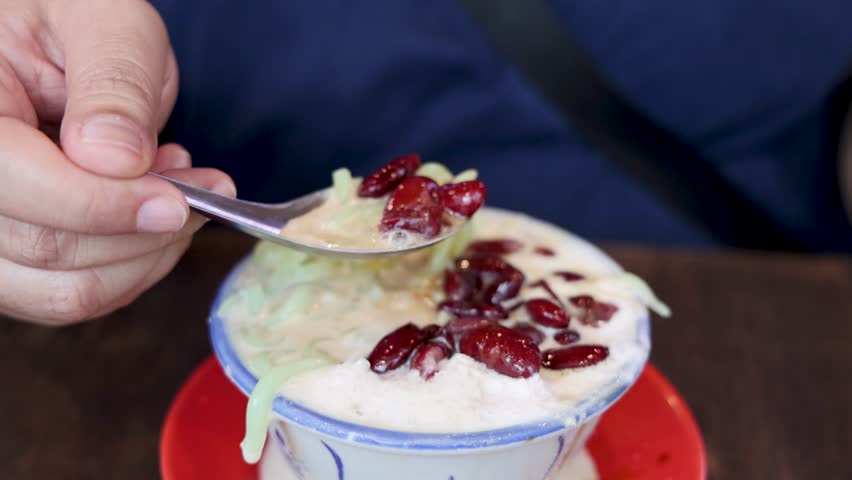 Close-up of hand scooping cendol dessert with red beans in a cozy indoor setting.
