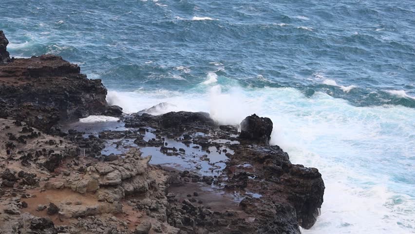 Slow motion video shows powerful ocean waves crashing against rugged volcanic rocks on the coastline near Kapalua, Maui, Hawaii, with mist rising and turquoise water swirling dramatically.
