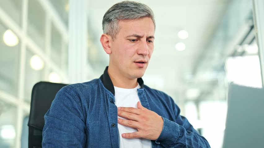 Worried man, dressed in a blue denim shirt and white t-shirt, experiences sharp chest pain in a modern office. His distressed expression indicates discomfort and concern for heart health.