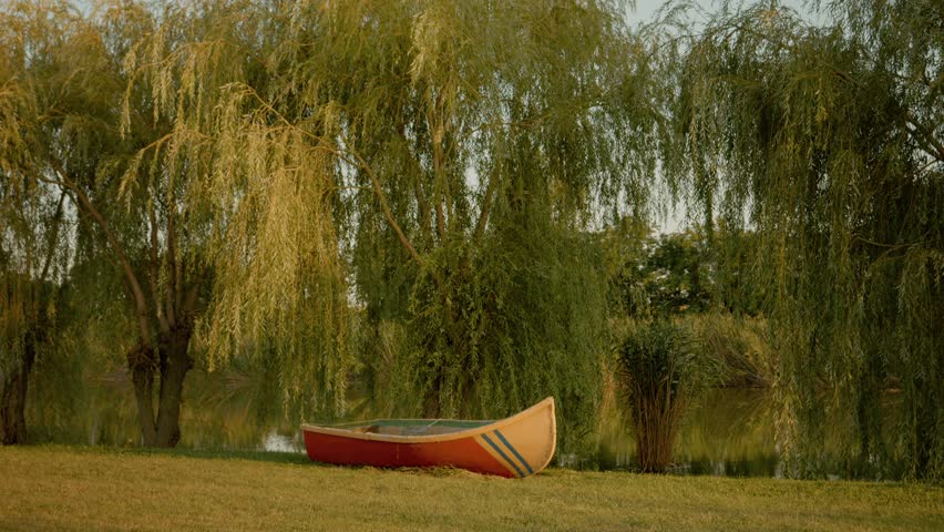 A colorful boat rests quietly on the grassy shore of a tranquil lake. Surrounding trees gently sway in the evening breeze as the sun sets on a peaceful scene.