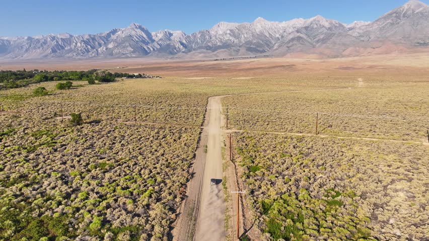 Drone tracks a car driving along a dusty dirt road through sagebrush flats in Owens Valley near Bishop California. The Eastern Sierra fills the horizon under clear summer light