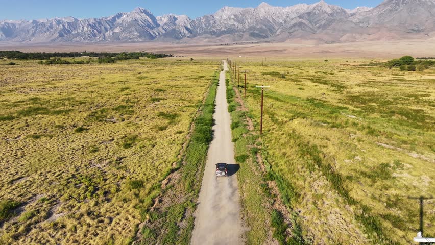 Drone tracks a car driving along a straight dirt road lined with power poles across open Owens Valley near Bishop California. Eastern Sierra peaks fill the horizon in clear summer light.