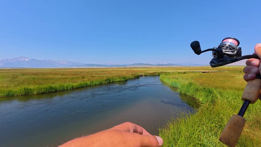 First-person shot of an angler casting a spinning rod into a clear, meandering river bordered by lush green grasses. Wide open Owens Valley landscape with the Sierra Nevada mountains on the horizon.