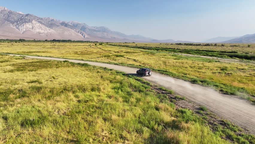 Drone pushes in tracks orbits a car driving along a dirt road in Owens Valley near Bishop California. Golden grassland and the Eastern Sierra mountains fill the background in warm clear daylight