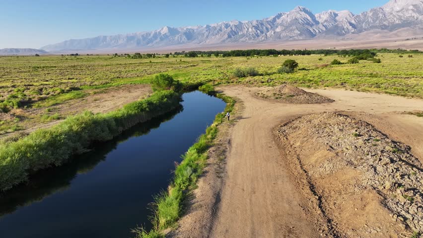 Drone orbits a man walking along a calm river channel on the Owens Valley floor near Bishop California with the Eastern Sierra in the background. Clear summer daylight dirt tracks and green bank edges