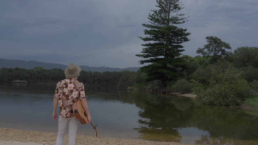 A still, rear-view shot of a man holding an acoustic guitar while standing or sitting at the edge of a calm lake. Captured from behind, the image conveys a peaceful, introspective mood.