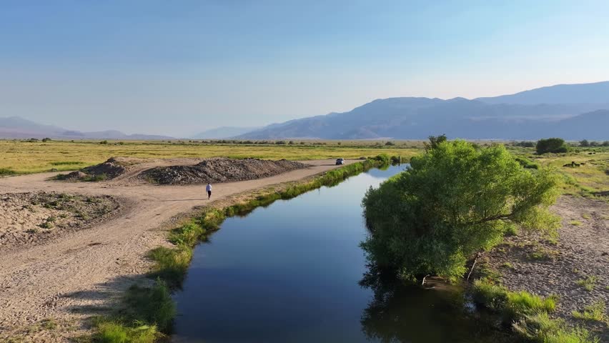 Drone begins in a close view of a man on a riverbank near Bishop California then ascends and pulls back to reveal a long canal the open Owens Valley and the Eastern Sierra in warm late day light.