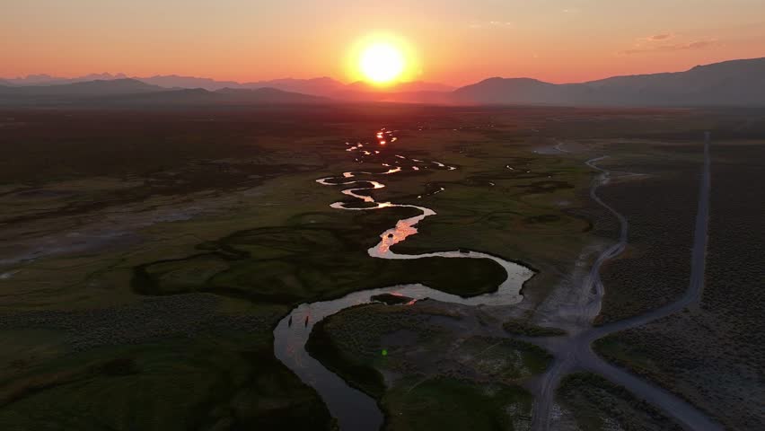Drone pushes in toward the setting sun over the meandering Owens River near Mammoth Lakes California. Golden hour light glows on S curve channels and open valley with distant Sierra peaks.