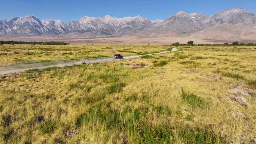 Drone tracks a car along a winding dirt road near Bishop California then ascends to reveal wide Owens Valley grasslands and the Eastern Sierra backdrop in clear summer light.