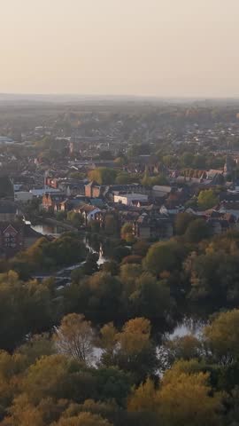 Vertical aerial drone footage over Ware, Hertfordshire, England, showing the River Lee, autumn woods, and town houses in warm golden light with a hazy afternoon sky.