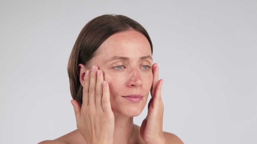 Portrait of a woman gently touching her face skin while applying face serum or facial oil on a light background. Hydrating skincare treatment and self-care concept with copy space.