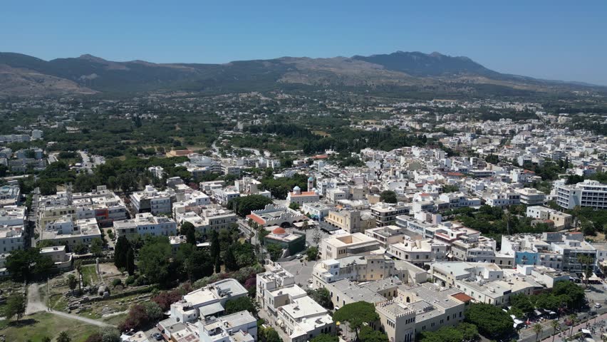 Aerial view of the sprawling white buildings of Kos city, nestled against the backdrop of rugged mountains and lush greenery, Kos, Greece.