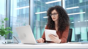Multitasking businesswoman works on digital tablet and laptop computer while sitting at desk at workplace in modern business office. Thoughtful female manager is engaged in a project, busy with tasks - Powered by Shutterstock - Get 15% off with code: PIKWIZARD15