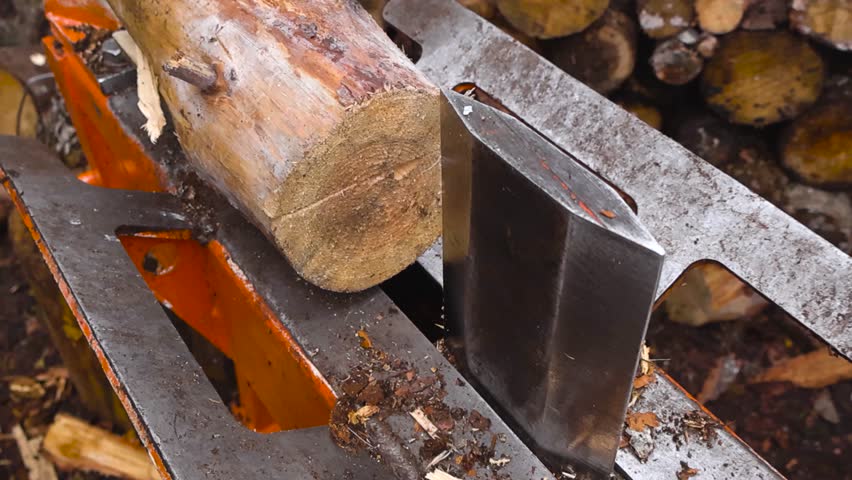 Close up view of a cut timber wooden pine log being split in pieces by a firewood splitting machine outdoors while cut wooden logs are in the vokeh blurry background at day time.Sawdust flying around.